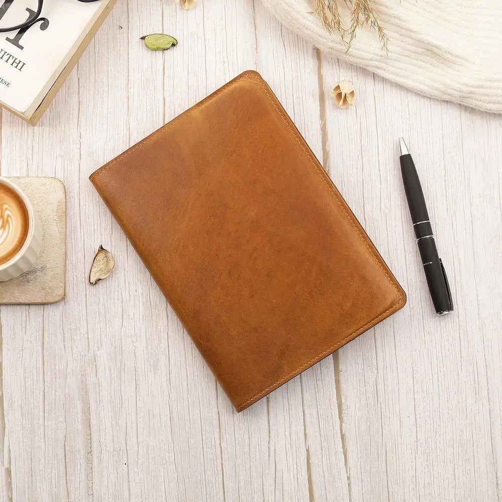 Tan leather diary cover  on a light wooden surface with a pen and coffee cup.