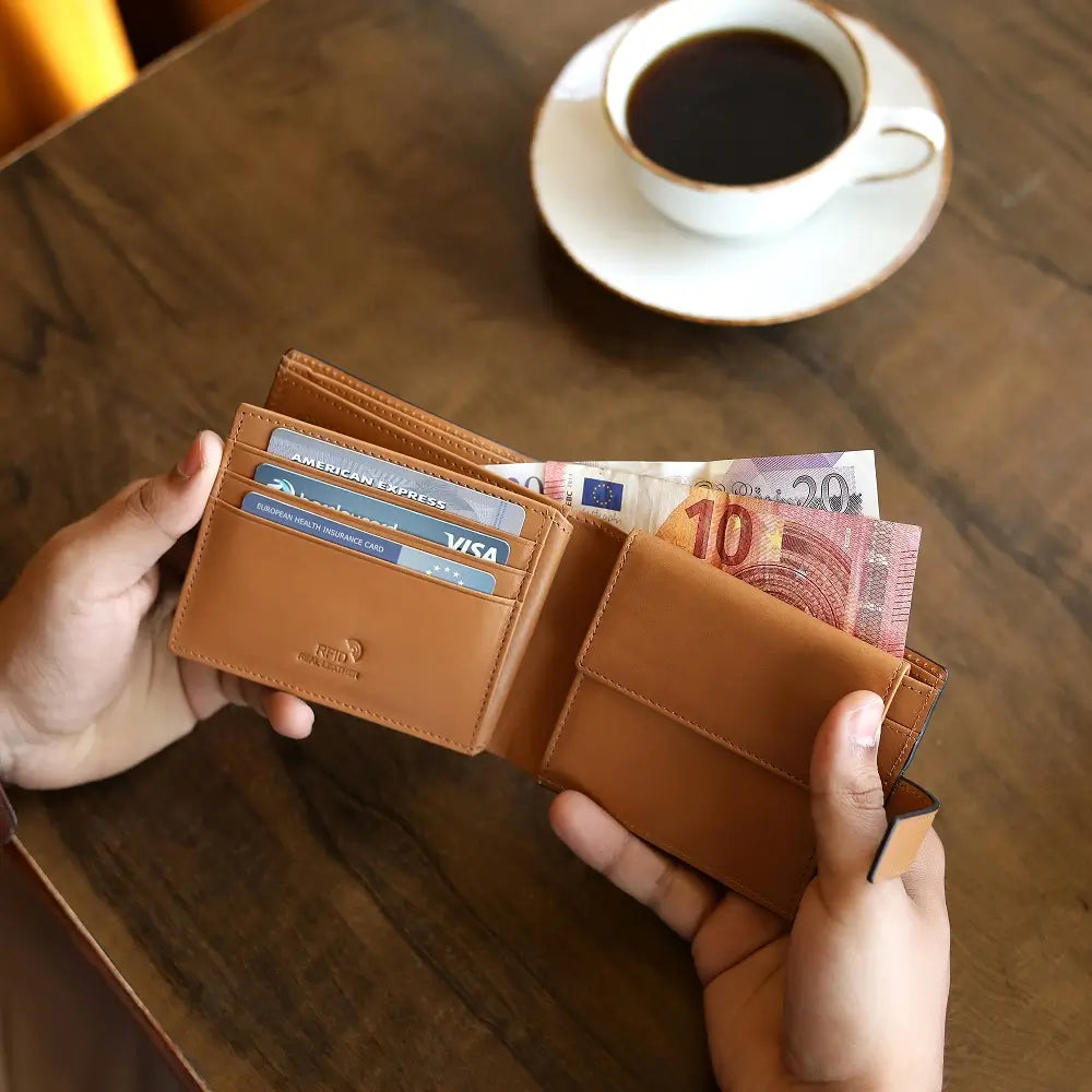 Tan leather wallet with money and cards held by hands on a wooden table with a cup of coffee.