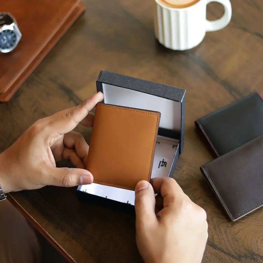 Person holding a tan leather wallet in an open box on a wooden table with other wallets and a cup in the background.