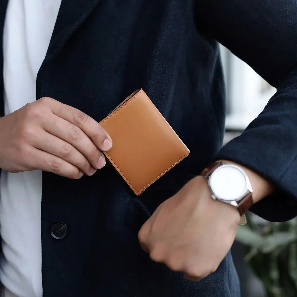 Person holding a tan leather wallet with a watch on the wrist, wearing a dark coat.