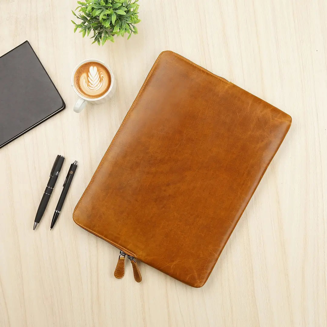 Tan leather laptop sleeve on a wooden surface with a notebook, pens, and coffee cup.