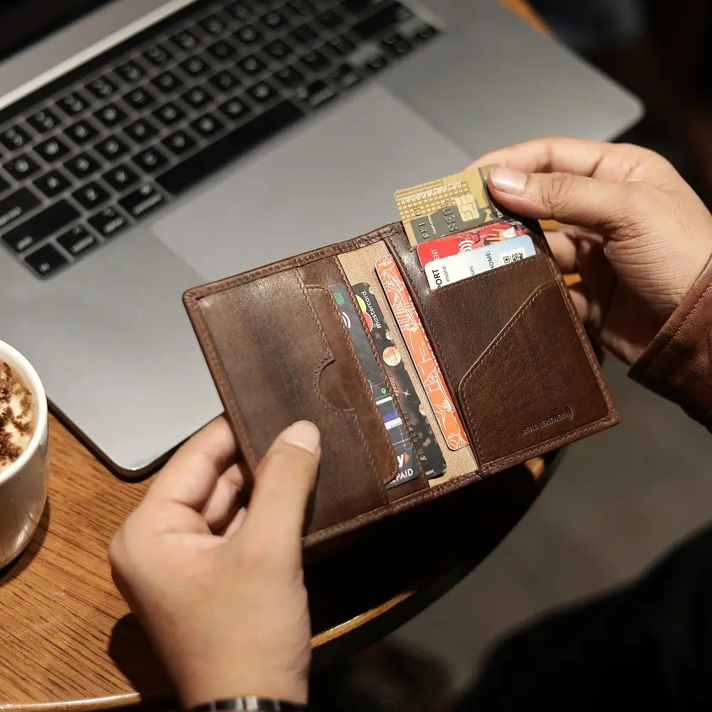 Person holding a brown leather wallet with cards in front of a laptop on a wooden table.