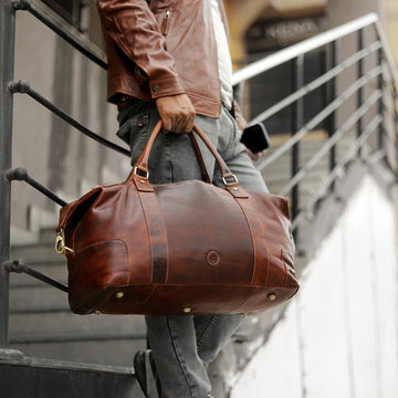 Man carrying Orion large full-grain tan leather holdall travel bag by Primehide outdoors, showing size and craftsmanship.