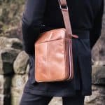 a man wearing a crossbody leather bag. the bag has a stripe down the side and a leather strap with a metal loop. The bag looks soft to touch with a luxurious feel