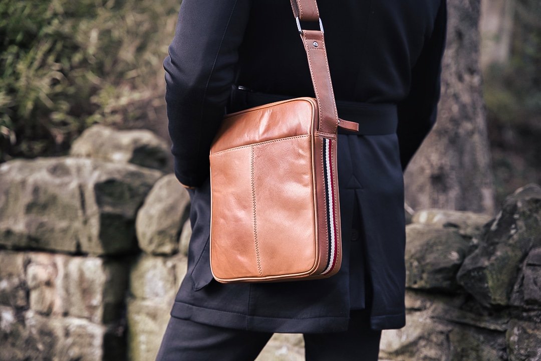 a man wearing a crossbody leather bag. the bag has a stripe down the side and a leather strap with a metal loop. The bag looks soft to touch with a luxurious feel