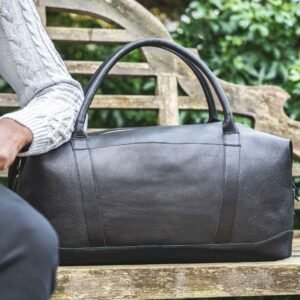 Man sitting stylishly next to a sleek black leather holdall, showcasing elegance and sophistication in travel accessories