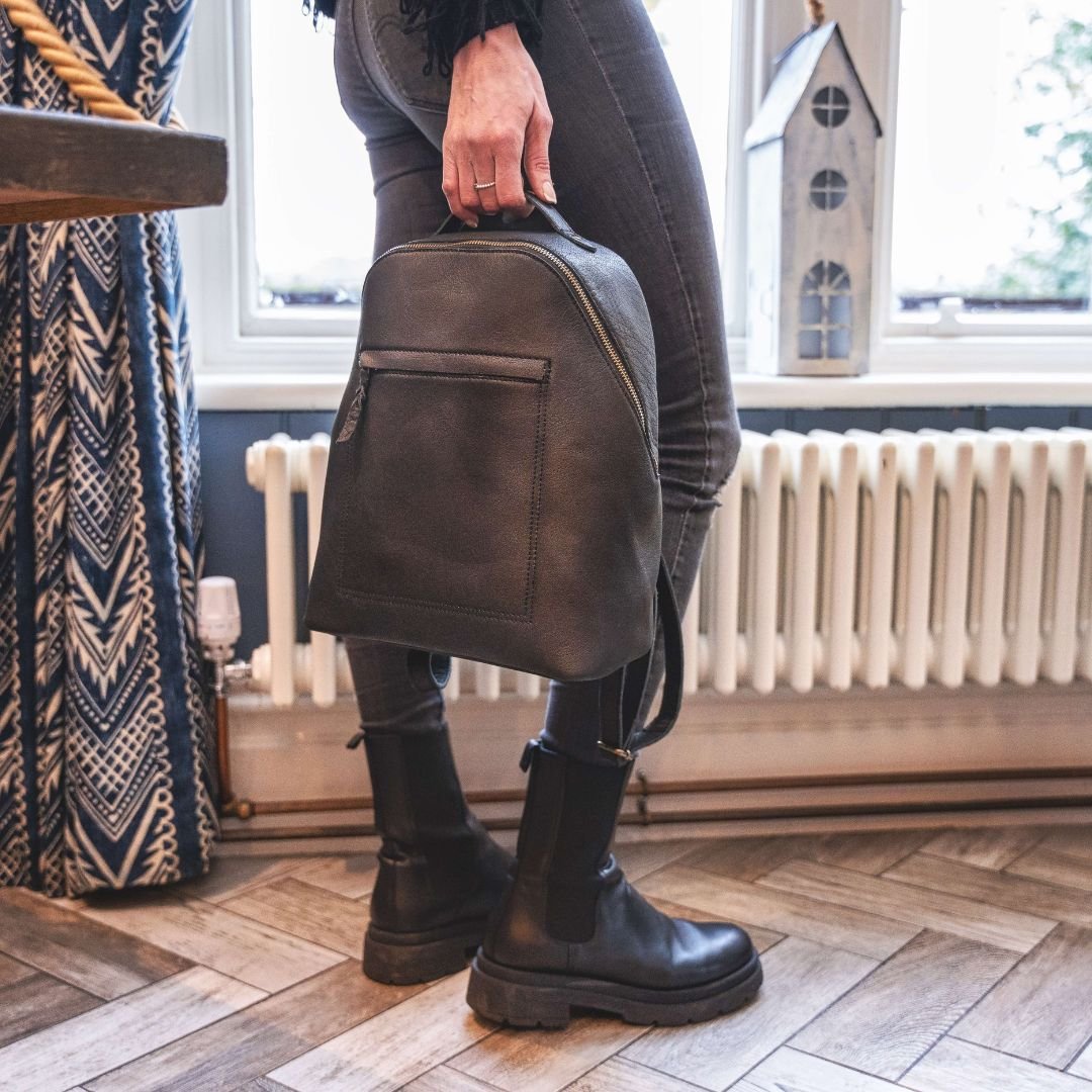 A woman standing in a bistro, holding a stylish black leather backpack, adding a touch of elegance to the casual ambiance.