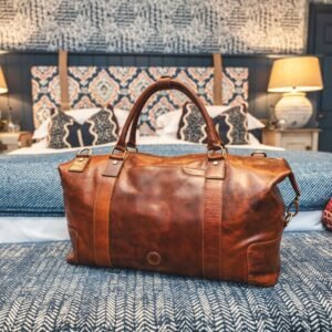 A luxurious brown leather weekend bag is elegantly placed on a neatly made hotel bed.