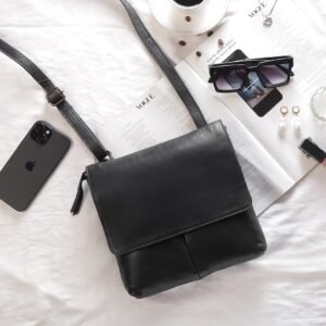 A black PRIMEHIDE crossbody organizer bag made of real leather laid out on a white surface, surrounded by a phone, sunglasses, a magazine, jewellry, and other accessories.