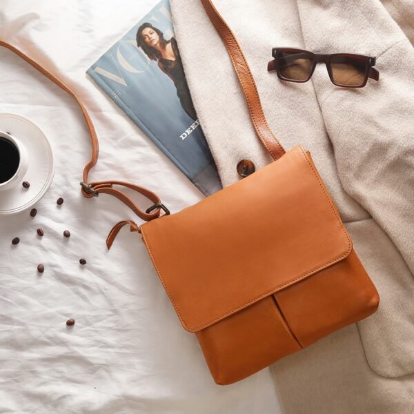 A cognac PRIMEHIDE crossbody organiser bag made of real leather laid out on a white surface, surrounded by a magazine, sunglasses, a cup of coffee, and other small items.