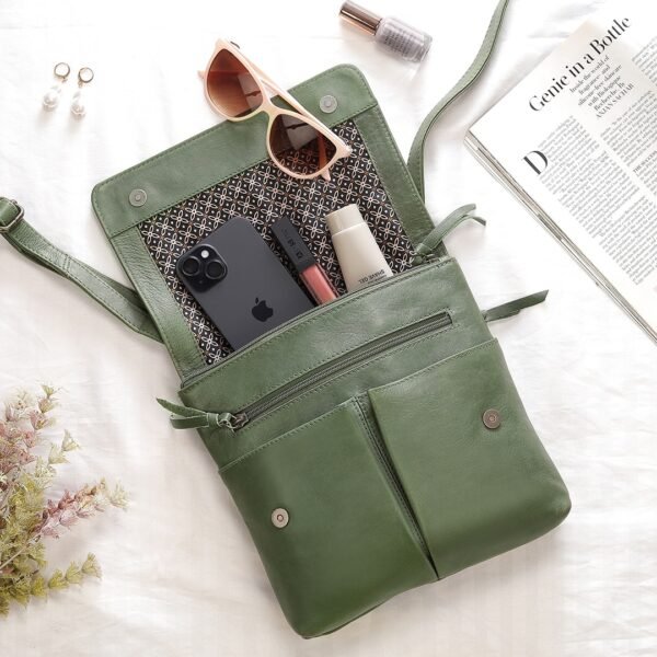 A green PRIMEHIDE crossbody organiser bag made of real leather laid out on a white surface, with its compartments open, showing a phone, sunglasses, makeup, and other items.