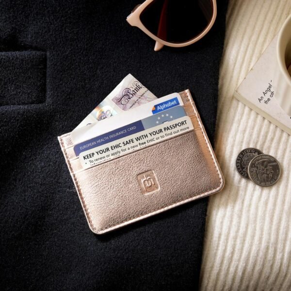 Rose gold leather card holder placed on a black coat with sunglasses, coffee, coins, and an EHIC card visible.